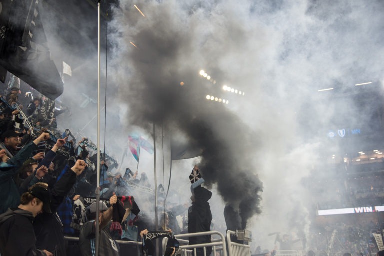 Dark Clouds and other fans cheering on Minnesota United while a plume of gray smoke drifts upwards.